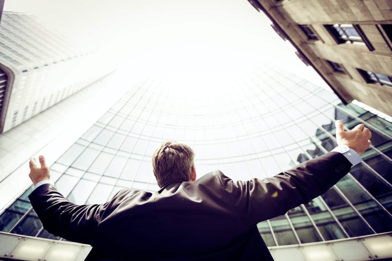 A man in a suit looking up at a skyscraper with arms open wide.