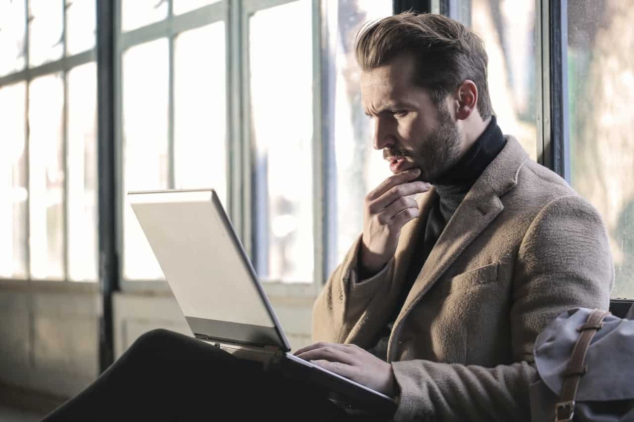 A man with a laptop on his lap and hand on his chin with a curious look on his face.