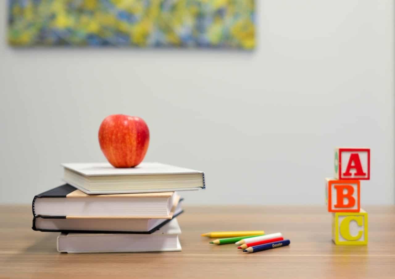 A stack of books with a red apple on top, some colored pencils and blocks with the letters "A", "B", and "C" sitting on a desk.