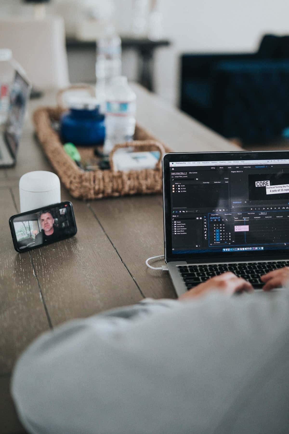 A man sitting at a dining room table typing on a laptop and using facetime on his cell phone.