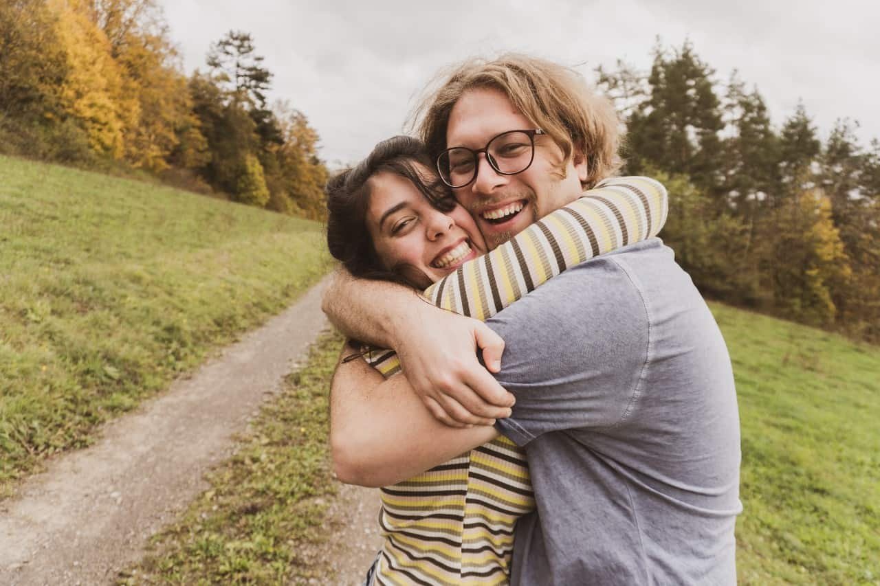 A man and woman hugging next to a road near a wooded area.