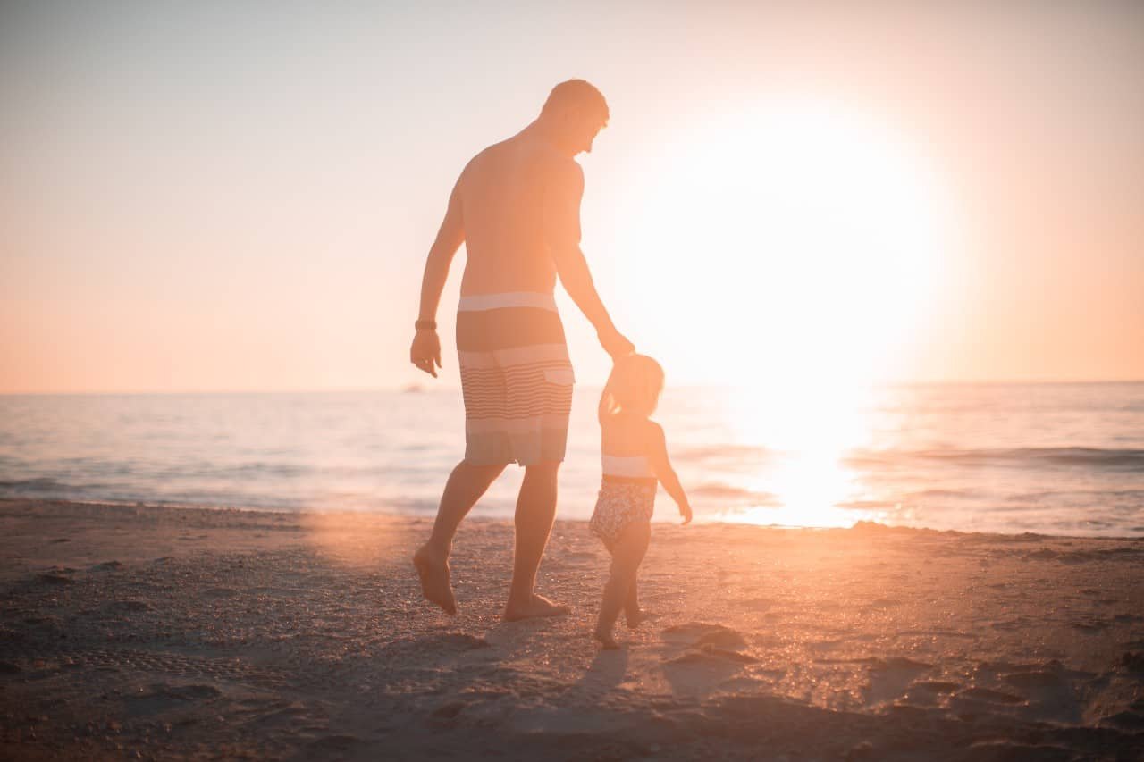 A father and his young daughter holding hands on the beach waking towards the water at sunset.