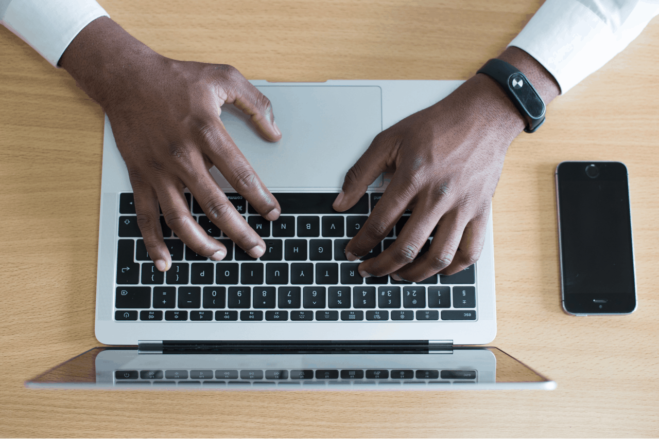 A man typing on a laptop with an iphone sitting next to it.
